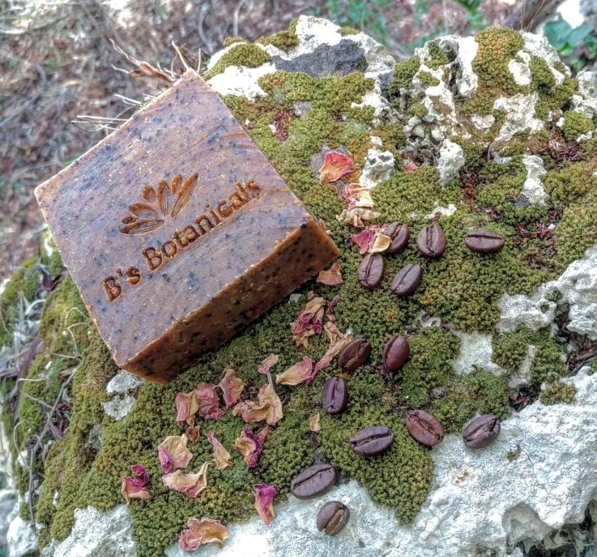 Bar of soap with 'B's Botanicals' branding on a mossy rock with coffee beans and dried flowers.
